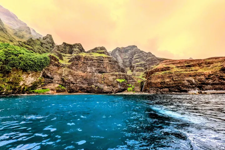 Rugged cliffs with lush greenery by a calm blue ocean under an orange sky.